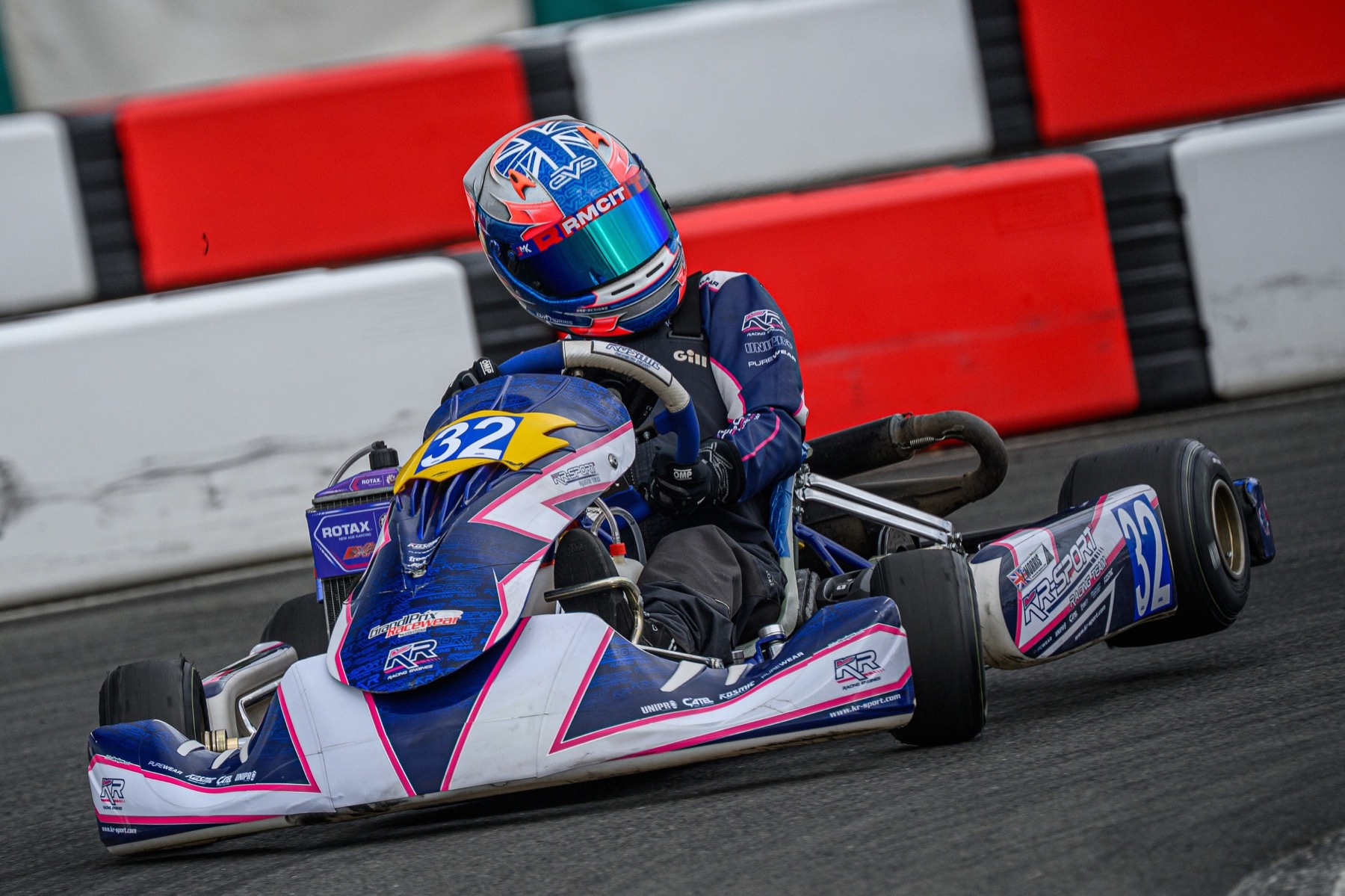 Eva sliding her navy, pink and white #32 kart through a corner, Union flag helmet on, red and white barriers behind